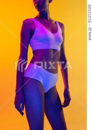 Cropped image of sportive, muscular built female body in white underwear isolated over yellow studio background in neon light 91352509