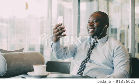 African American businessman in formal clothes smiling, laughing and talking to family through smartphone camera in cafe. He waves his hand and gesticulates happily with earbuds in ears 91356025