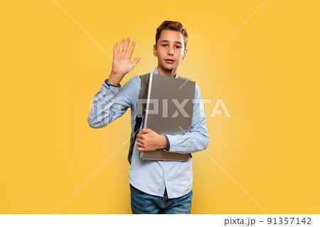 Concept of modern technologies and education. A friendly young Caucasian boy in a blue shirt and backpack, holding a laptop and waving. Yellow background. Copy space 91357142