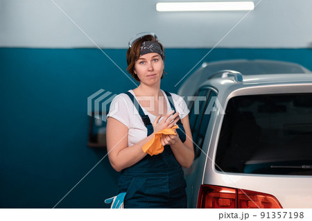 Portrait of a caucasian young woman in a blue coveralls waping her hands with a rag. Car and workshop in the background. Work in an auto repair shop and garage Portrait of a caucasian young woman in a blue coveralls waping her hands with a rag. Car and workshop in the background. Work in an auto repair shop and garage 91357198