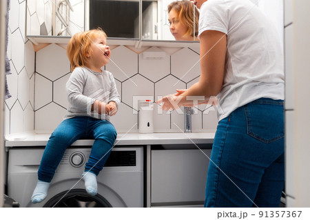 Happy little girl sitting on a countertop and looks to mother how she washes hands. Bedtime hygiene procedures Happy little girl sitting on a countertop and looks to mother how she washes hands. Bedtime hygiene procedures 91357367