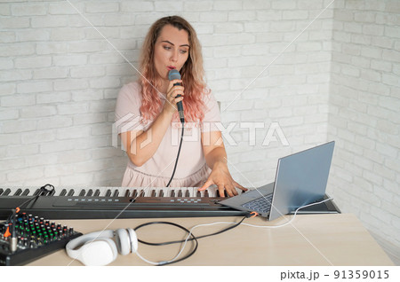 A woman records a vocal lesson using a laptop and accompanying on a keyboard while at home. The teacher sings a song into the microphone and plays the electronic piano. A blogger is recording a video. 91359015