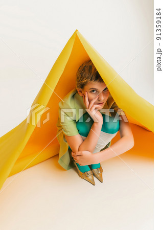 Stylish young girl posing in light green suit, hiding under big yellow paper piece isolated over grey studio background 91359184