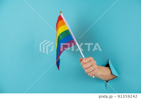 A woman's hand sticks out of a hole in a blue paper background and holds an lgbt flag. 91359242