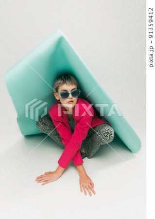 Portrait of young girl in bright jacket and vintage pants posing, sitting under big paper piece isolated over grey studio background. Retro 91359412