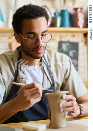 Serious guy in eyeglasses and apron using handtool while creating clay jug 91359892