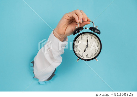 A woman's hand sticks out of a hole in a blue paper background and holds an alarm clock. A woman's hand sticks out of a hole in a blue paper background and holds an alarm clock. 91360328