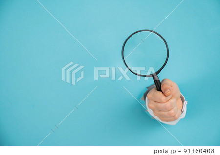 A woman's hand sticking out of a hole in a paper blue background holds a magnifying loupe. Copy space. A woman's hand sticking out of a hole in a paper blue background holds a magnifying loupe. Copy space. 91360408