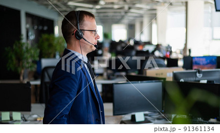 Portrait of a male call center operator in headset at workplace. A man works in an open space office answering customer calls. Support service or hotline. Agent of a telemarketing company. Portrait of a male call center operator in headset at workplace. A man works in an open space office answering customer calls. Support service or hotline. Agent of a telemarketing company. 91361134