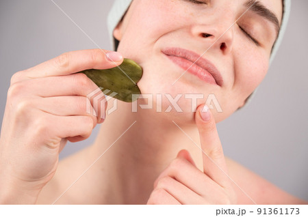 Portrait of a young woman massages her face with a gouache scraper on a white background. Portrait of a young woman massages her face with a gouache scraper on a white background. 91361173