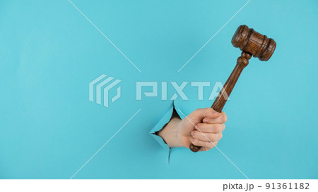 A woman's hand with a wooden judge's gavel sticks out of a hole in a blue background. Copy space. A woman's hand with a wooden judge's gavel sticks out of a hole in a blue background. Copy space. 91361182