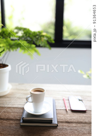 White coffee cup and phone with notebooks and plant pot on brown wooden table in front of window White coffee cup and phone with notebooks and plant pot on brown wooden table in front of window 91364653