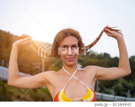 Smiling girl in a bikini holding her pigtails while standing against the backdrop of a green landscape on a summer day. Beach vacation. Summer weekend Smiling girl in a bikini holding her pigtails while standing against the backdrop of a green landscape on a summer day. Beach vacation. Summer weekend 91364842