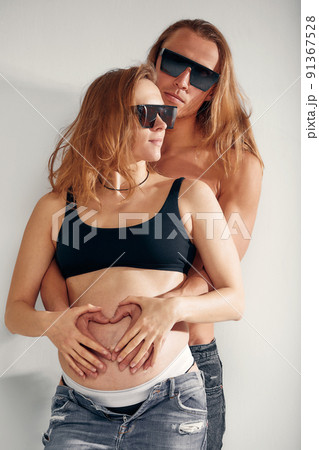 husband holding belly of his pregnant wife wearing sunglasses making symbol heart hands. Pregnant woman and loving handsome man hugging tummy at home. Loving Couple. Parenthood concept. Baby Shower. husband holding belly of his pregnant wife wearing sunglasses making symbol heart hands. Pregnant woman and loving handsome man hugging tummy at home. Loving Couple. Parenthood concept. Baby Shower. 91367528