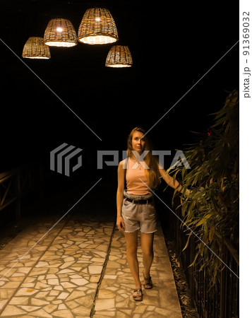 A woman stands on a marble path in a park bamboo alley at night, illuminated by lamps with wicker lampshades. Evening walk around the southern arboretum. Focus on the girl A woman stands on a marble path in a park bamboo alley at night, illuminated by lamps with wicker lampshades. Evening walk around the southern arboretum. Focus on the girl 91369032