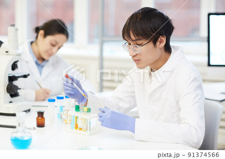 Serious young Asian lab technician in protective gloves and white coat sitting at table in laboratory and using pipette while adding reagent into test tube 91374566