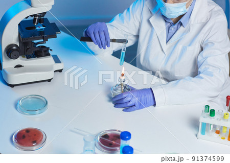Close-up of unrecognizable lab technician in gloves and mask sitting at desk and using burner to catalyze chemical reaction Close-up of unrecognizable lab technician in gloves and mask sitting at desk and using burner to catalyze chemical reaction 91374599