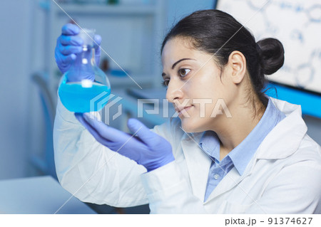 Serious young Indian female chemist in lab coat and gloves holding beaker and analyzing color of chemical liquid Serious young Indian female chemist in lab coat and gloves holding beaker and analyzing color of chemical liquid 91374627