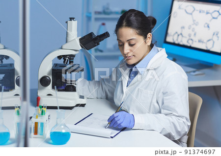 Serious busy young mixed race chemist in white coat sitting at desk with microscope and making notes in clipboard while doing laboratory research Serious busy young mixed race chemist in white coat sitting at desk with microscope and making notes in clipboard while doing laboratory research 91374697