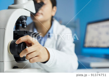 Close-up of female scientist in lab coat sitting at table and using adjustment knob of microscope Close-up of female scientist in lab coat sitting at table and using adjustment knob of microscope 91374751