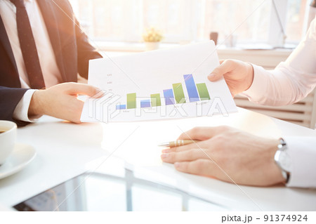 Close-up of unrecognizable businessman sitting at table in office and giving sales report with graph picture to colleague 91374924