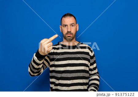 Hispanic man with beard wearing striped sweater standing over isolated blue background Showing middle finger, impolite and rude fuck expression. 91375122
