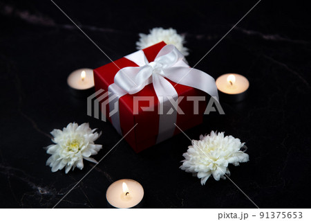 In the center of the black marble is a gift to a red box with a white ribbon surrounded by lighted candles and white flowers. High quality photo 91375653