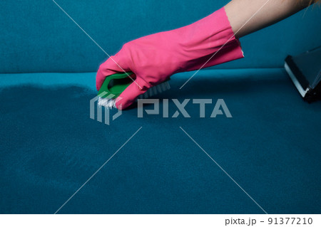 Dry cleaner's employee hand in protective gloves removing stain from gray sofa with brush and special foam. Early spring regular cleanup. Commercial cleaning company. Closeup. Top view. Dry cleaner's employee hand in protective gloves removing stain from gray sofa with brush and special foam. Early spring regular cleanup. Commercial cleaning company. Closeup. Top view. 91377210
