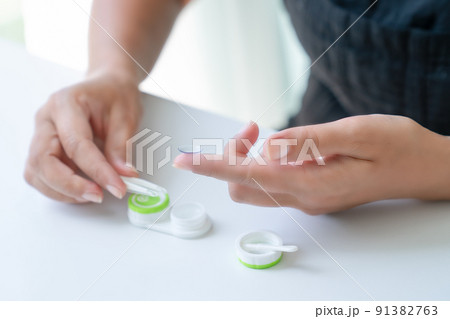 Woman puts it down contact lens on her index finger with tweezers. There is container for lenses nearby. Close-up, side view Woman puts it down contact lens on her index finger with tweezers. There is container for lenses nearby. Close-up, side view 91382763