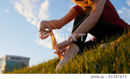 Young woman put on her white pointe shoes and ties it up - sitting on the green grass at sunset 91387624