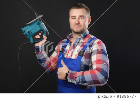 Portrait of a caucasian male repairman in a work uniform with an electric jigsaw in your hand Showing thumbs up. Studio portrait of an artisan businessman 91396777