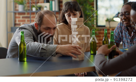 Positive people having fun with society game in living room, playing with wooden tower blocks. Men and women enjoying game with building square pieces on structure for entertainment. Positive people having fun with society game in living room, playing with wooden tower blocks. Men and women enjoying game with building square pieces on structure for entertainment. 91398272