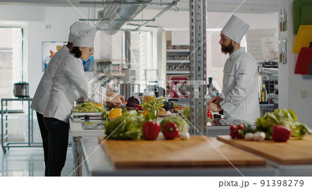 Diverse team of cooks preparing organic ingredients for cuisine dish, making delicious gourmet meal with salad and vegetables in professional kitchen. Man and woman cooking food. Tripod shot. 91398279