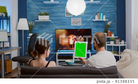 Young woman sitting next to boyfriend holding vertical digital tablet with green screen in online conference or group video call. Couple using touchscreen device with chroma key watching vlog. Young woman sitting next to boyfriend holding vertical digital tablet with green screen in online conference or group video call. Couple using touchscreen device with chroma key watching vlog. 91398335