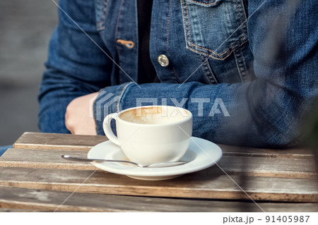 Person in jean jacket sits in outdoor cafe with empty cup of coffee on table top 91405987