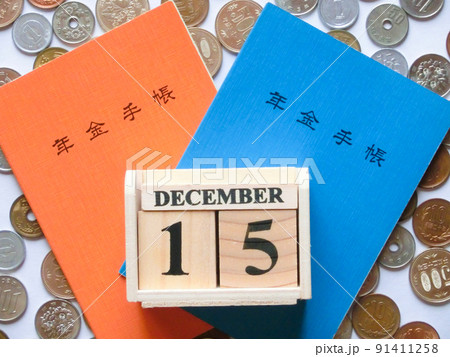 散らばった日本の硬貨を背景に重なった橙色と青色の年金手帳と12月の年金支給日を表した木製カレンダー 散らばった日本の硬貨を背景に重なった橙色と青色の年金手帳と12月の年金支給日を表した木製カレンダー 91411258