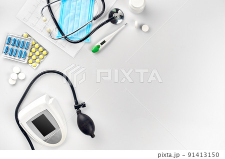 White electric tonometer with stethoscope, multicolored pills, mask and thermometer on white table. Top view 91413150