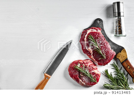 Two fresh raw steaks with herbs, garlic, olive oil, pepper, salt, rosemary and a kitchen ax on white background. Top view. 91413465