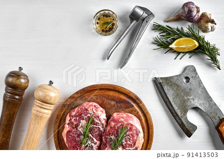 Two raw beef steak with spices on wooden board and kitchen ax on white background, top view Two raw beef steak with spices on wooden board and kitchen ax on white background, top view 91413503