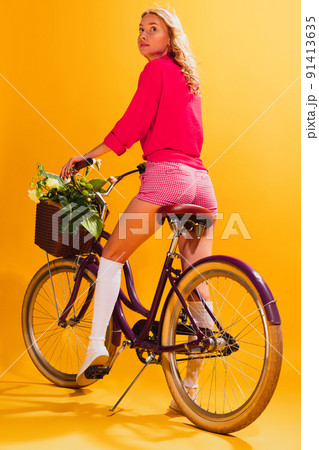 Portrait of beautiful young girl in pink outfit posing with retro bike isolated over yellow studio background. Cheerful lifestyle 91413635