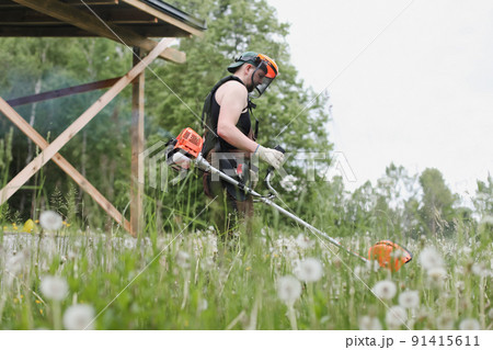 Man mowing tall grass with petrol lawn trimmer in the garden or backyard. Process of lawn trimming with hand mower 91415611