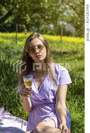 Happy beautiful girl on a picnic in nature. Young girl in a purple dress having a picnic with a glass of champagne or white wine. Summer time Happy beautiful girl on a picnic in nature. Young girl in a purple dress having a picnic with a glass of champagne or white wine. Summer time 91416349