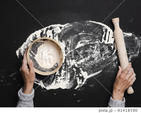 Female hands hold a round wooden sieve with white wheat flour on a black table, top view Female hands hold a round wooden sieve with white wheat flour on a black table, top view 91418590
