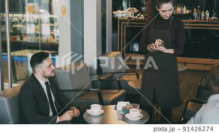 Two cheerful multi-ethnic businessmen in formal clothes sitting, smiling and making order in modern cafe during lunch break. Young and optimistic waitress writing down their dishes Two cheerful multi-ethnic businessmen in formal clothes sitting, smiling and making order in modern cafe during lunch break. Young and optimistic waitress writing down their dishes 91426274