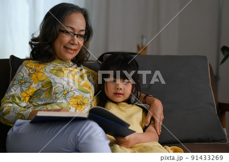 Happy moments with grandmother and little granddaughter reading book together on sofa Happy moments with grandmother and little granddaughter reading book together on sofa 91433269