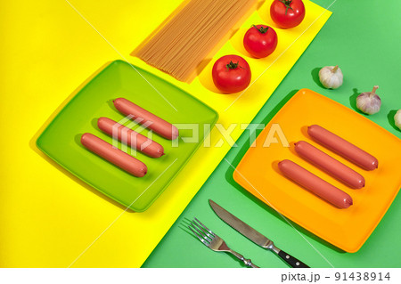 A lot of raw sausages on plate. On green and yellow background with pasta and vegetables, top view. A lot of raw sausages on plate. On green and yellow background with pasta and vegetables, top view. 91438914