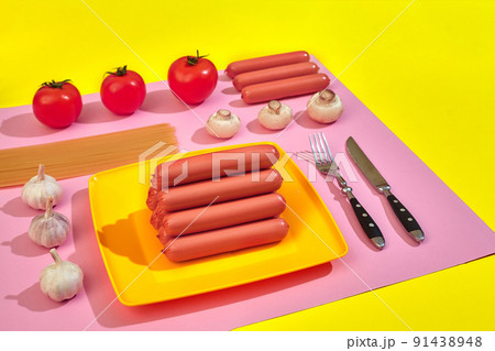 A lot of raw sausages on plate. On pink and yellow background with pasta and vegetables, top view. 91438948