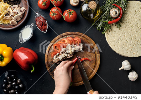 Female hand cut chilli, mushrooms and tomatoes on wooden board on kitchen table, around lie ingredients for pizza: vegetables, cheese and spices. Female hand cut chilli, mushrooms and tomatoes on wooden board on kitchen table, around lie ingredients for pizza: vegetables, cheese and spices. 91440345