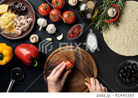 Female hand cut tomatoes on wooden board on kitchen table, around lie ingredients for pizza: vegetables, cheese and spices. 91440385