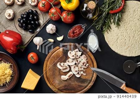 Cutting wooden board with traditional pizza preparation ingredients: cheese, tomatoes sauce, mushrooms, olive oil, pepper, spices. Black texture table background 91440439